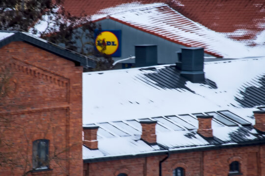 Polska, Ostrowiec Świętokrzyski, 31 grudnia 2025, 14:59. Szyld sklepu LIDL &ndash; dyskont spożywczy &ndash; ponad dachami zimą. Szyld przedstawia rozpoznawalne niebiesko-ż&oacute;łto-czerwone logo na szarej fasadzie. 