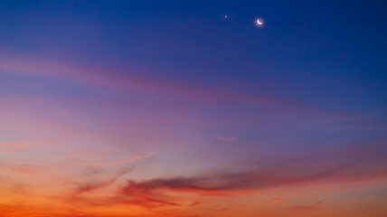 Obraz premium Crescent Moon and Planets in the Twilight Sky Over Soft Sunset Clouds,A breathtaking view of a thin crescent moon shining alongside bright planets in a deep blue twilight sky. The lower horizon.