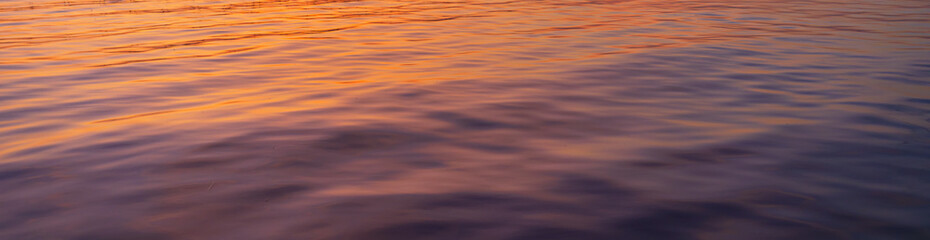 Golden Hour Reflections on Calm Water Surface,Abstract nature background of gentle ripples on a water surface during sunset, featuring a vibrant blend of golden orange and deep purple hues. © banjongseal324