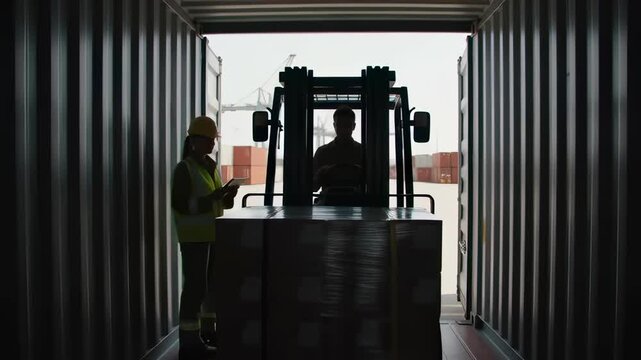 Logistics supervisor checking cargo on a tablet as a forklift loads a pallet into a shipping container at a sunny port, illustrating supply chain efficiency.