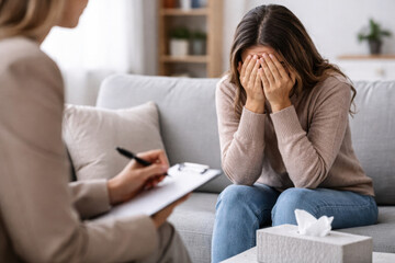 Woman covering her face during psychotherapy session, sitting on sofa opposite therapist. Concept of psychological help, emotional crisis, stress, support and mental health.