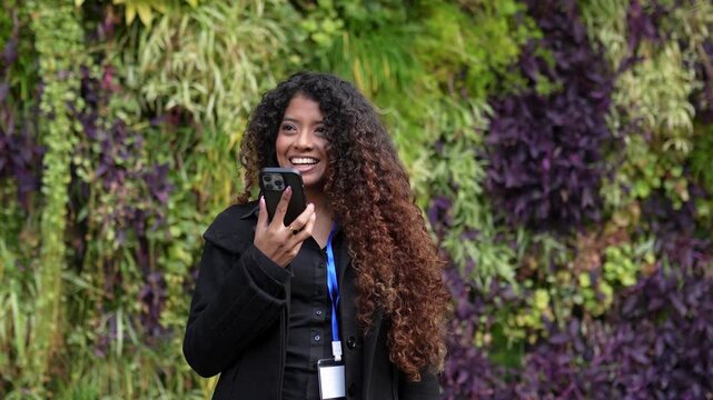Smiling businesswoman with long curly hair using her mobile phone to send a voice message. She stands in front of a green wall of plants