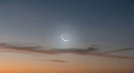 Crescent moon shining brightly above soft orange and blue clouds at sunset creating serene evening atmosphere for religious celebrations lunar observation and peaceful nature backgrounds for design