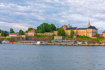 Obraz premium Akershus Fortress situated on headland overlooking the calm waters of Oslofjord in Oslo, Norway. Medieval fortress complex is surrounded by lush trees and fortified walls with boat dock at its base