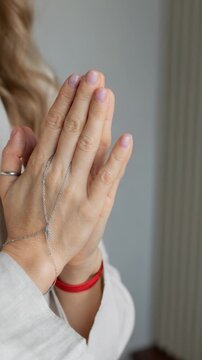Woman in a beige robe clasps their hands together in a prayerful pose, adorned with a delicate silver bracelet and a red string, sense of peace, reflection, and spirituality.