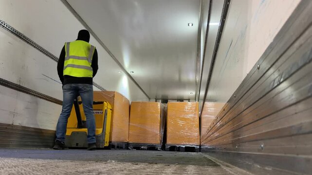 Warehouse operative unloading orange-wrapped pallets from a truck trailer using an electric ride-on pallet jack