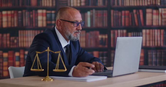Attorney consulting client via laptop in library of law office. Court cases paperwork and corporate agreement research, legal expertise by senior lawyer, contract review at desk with justice scales