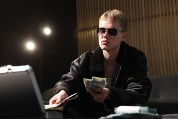 Young businessman in jacket and sunglasses putting money into metal briefcase at table in dark room