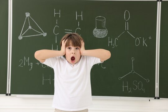 Shocked schoolboy near chalkboard with chemical formulas in classroom - Powered by Adobe