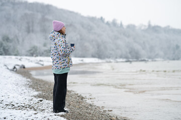 Girl with a cup of a hot drink stands on the snowy shore of a winter sea and looks into the distance. © MIRACLE MOMENTS