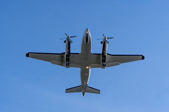 Small twin engine airplane with a dramatic outline and blue sky in the background flying overhead as it is coming in for a landing at an airport