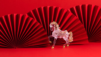 Year of the Horse 2026. A transparent horse figurine with gold details stands on a red background with decorative paper fans.