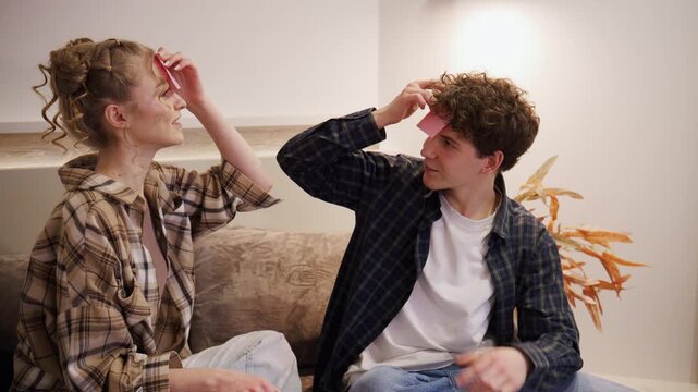 Two people play a guessing game with sticky notes on their foreheads in a room during daytime