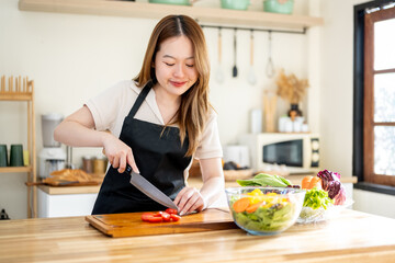 An Asian woman is slicing tomatoes on a kitchen table to make a salad. The imagery relates to healthy eating and dieting.