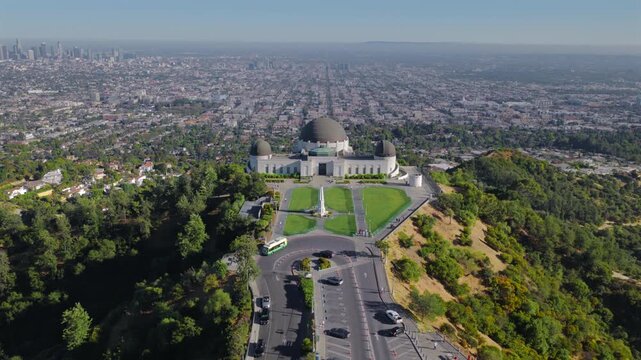 Aerial drone shot above Griffith Observatory parking road with cars, green hills and vast Los Angeles California urban sprawl stretching to the horizon. Downtown Los Angeles on a background.