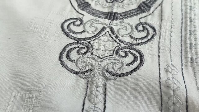 Close-up of grey embroidery on white fabric, featuring elegant scroll patterns and intricate stitch details on a traditional shirt