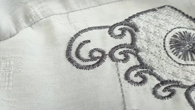 Close-up of grey embroidery on white fabric, featuring elegant scroll patterns and intricate stitch details on a traditional shirt