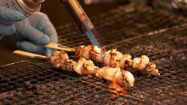 Chef using a blowtorch to cook fresh squid skewers at a Tokyo street stall.