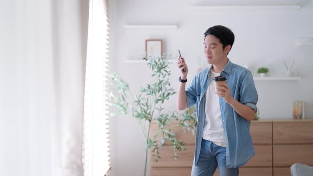 Happy Asian man drinking hot coffee in the morning using the phone while standing in the living room at home. Handsome young man walking around with a cup of hot drink, looks at a smartphone