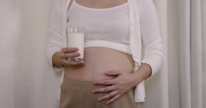 Closeup of asian pregnant belly with hand holding milk glass symbolizing nourishment, prenatal health, and maternal selfcare during quiet third trimester lifestyle at home