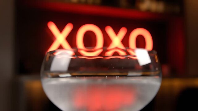 A close-up shot captures the clear glass of liquid reflecting the vibrant red xoxo neon sign with a romantic, moody atmosphere in a dark indoor setting.