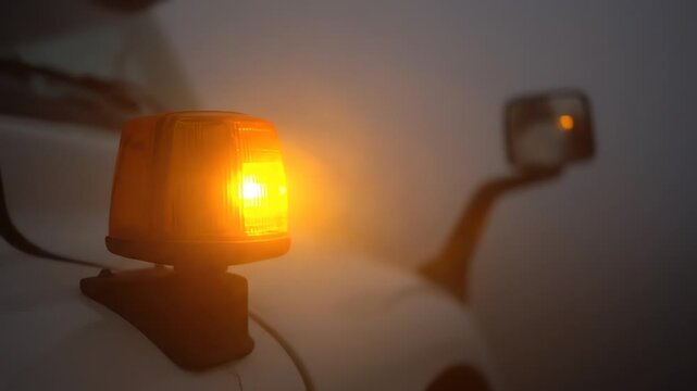 Captured in a close-up shot, the amber warning light flashes intensely, casting an urgent orange glow from a utility vehicle into the thick, mysterious fog.