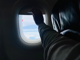A passenger's hand reaches up to close the window shade of a commercial aircraft during a flight to block out sunlight and rest.