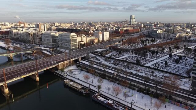 Aerial drone view of Paris suburban landscape covered in snow during winter, featuring railway infrastructure crossing river with urban buildings and architecture under overcast sky, Paris, France