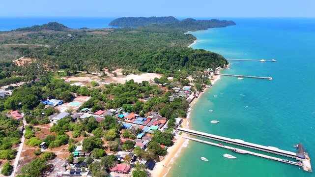 Aerial drone view of Koh Phayam village, showing the coastline, piers, boats, and a lush tropical landscape, surrounded by turquoise sea.