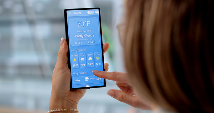 Young Woman Checking Weather Forecast On Smartphone App In Living Room