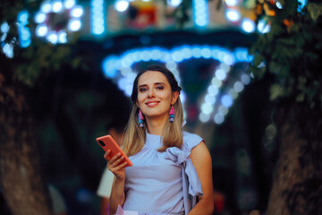 Smiling Woman Holding a Mobile Phone in an Amusement Park. Happy lady holds a mobile phone while...