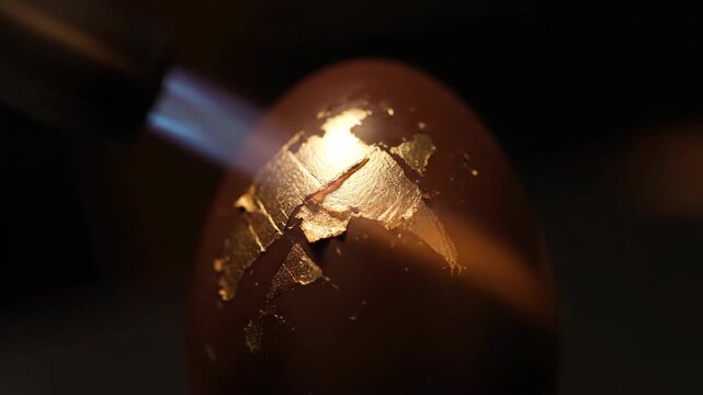 Conceptual macro of a blowtorch flame heating a brown egg adorned with a gold leaf world map. Symbolizes global warming, wealth under pressure, and fragile economic conditions.
