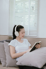 Obraz premium A young Asian woman sits on a sofa at home, wearing wireless headphones and smiling while using her smartphone, enjoying music and a relaxed, modern digital lifestyle.