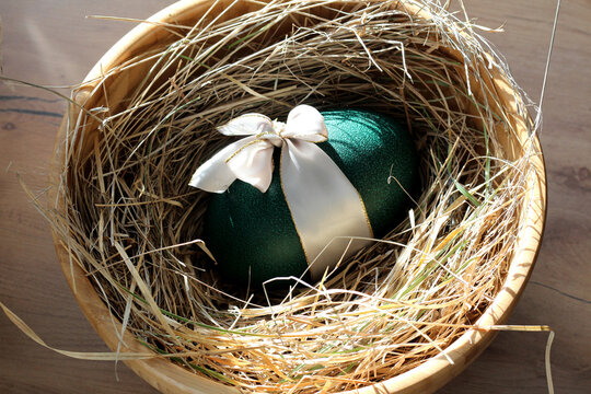 Emerald green decorative Easter emu egg with a silky white ribbon bow, nestled in a rustic hay nest inside a wooden bowl. Elegant holiday composition with natural textures and soft sunlight shadows.