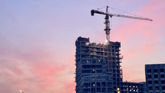 Construction site with towering cranes and multi-story residential buildings under development against a vibrant sunset or sunrise sky. Real estate investment and urban growth concept.
