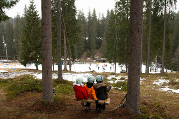 Children enjoying a scenic winter adventure while seated on a swing, overlooking a busy ski slope surrounded by tall coniferous trees