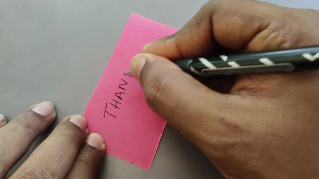 Indian or Asian person hand with pen writing thanks message or word on blank sticky note paper on a desk background. Expressing handwritten appreciation or gratefulness text card concept.