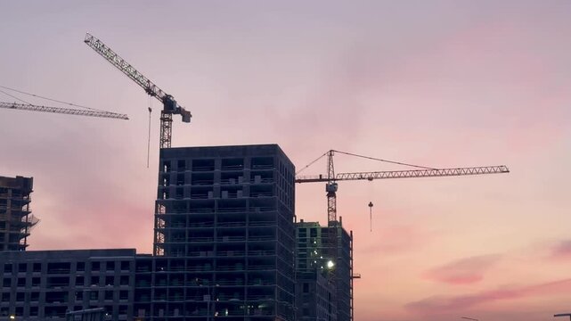 Construction site with towering cranes and multi-story residential buildings under development against a vibrant sunset or sunrise sky. Real estate investment and urban growth concept.
