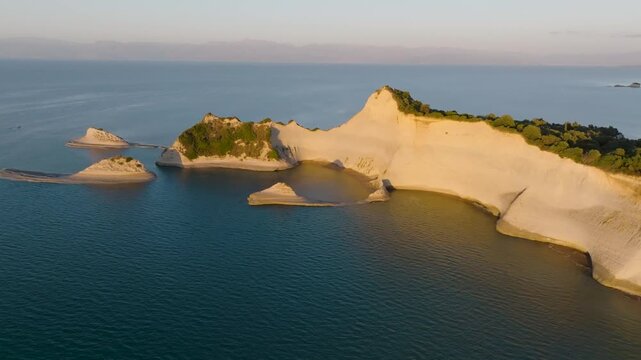 Slow aerial flight over the turquoise water towards the iconic white cliffs of Cape Drastis, Corfu. Cinematic drone view of the famous Greek island landmark at sunset