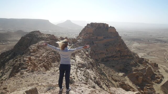 Tourist woman with blonde hair raises her arms while looking over Douiret, Tunisia, as wind blows across the hilltop, capturing freedom, travel inspiration, and the vast desert village landscape
