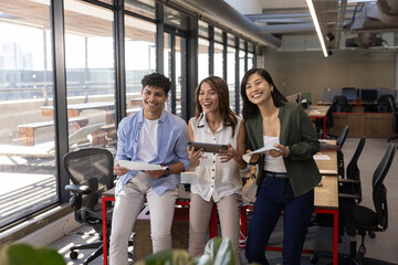 Diverse coworkers leaning against desk holding documents and tablet discussing in office