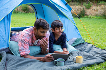 Indian father with son lying inside blue tent on backyard lawn watching tablet with enamel mugs © wavebreak3