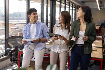Diverse coworkers smiling and discussing work on tablet and notebook at office desk