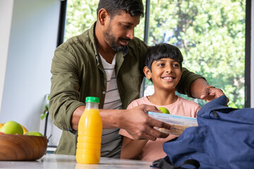 Indian father with son packing lunch at kitchen counter with plastic container, navy blue backpack © wavebreak3