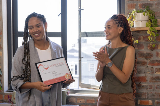 Diverse female coworkers celebrating framed certificate at office with window plant shelf
