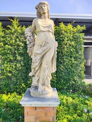 Obraz premium Classical Garden Statue of a Woman in Ancient Style Surrounded by Greenery A classical stone statue of a woman inspired by ancient Greek and Roman art, standing gracefully in a garden setting.