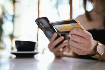 Close-up of a woman holding a credit card and smartphone in a café, making a mobile payment or online purchase, representing cashless transactions, digital banking, and modern lifestyle. © Worawi