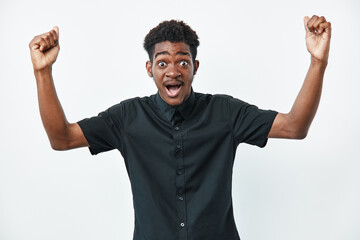Fototapeta premium Young black man with dark skin wearing black shirt raising arms and expressing excitement or surprise on white background. Portrait of emotional african male.