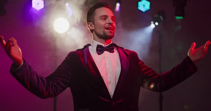 Showman presenter host emcee performer on stage. Tuxedo and bowtie shine under spotlight. Colorful lighting and smoke frame a welcoming gesture. Concept shows celebration and engagement.