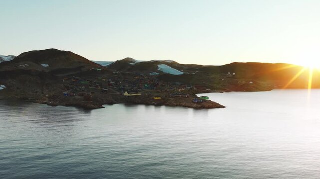 Camera orbits around Iqqortoormiit, Greenland revealing a morning sunrise washing golden light on the town.